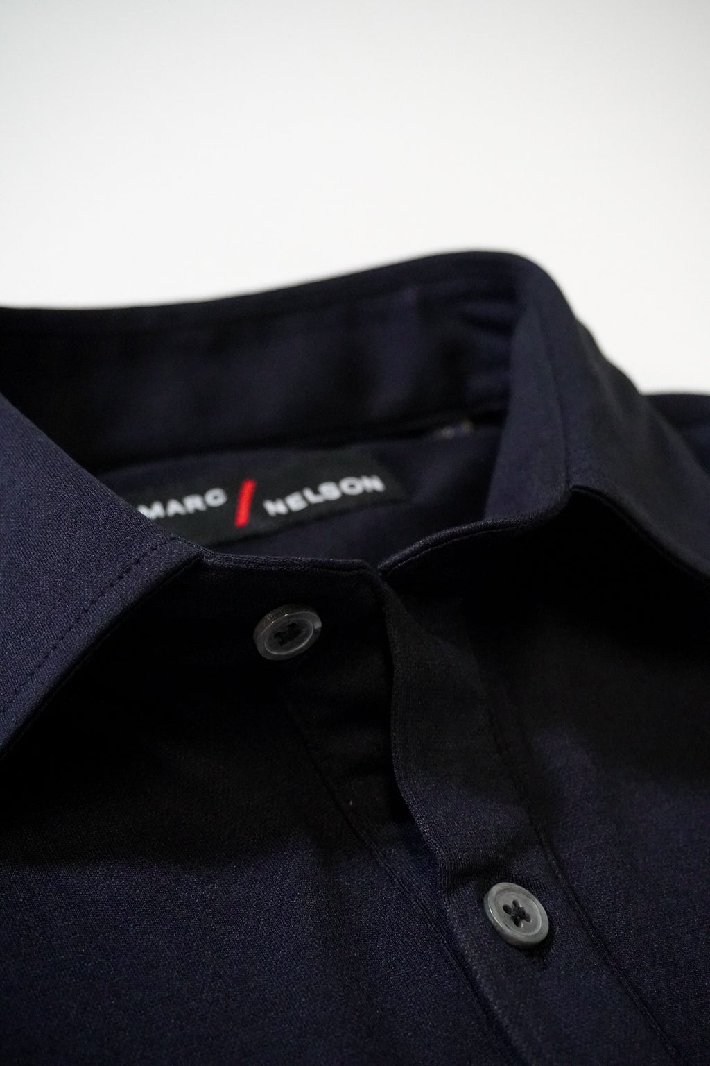 Close-up of a neatly folded dark navy blue collared shirt with gray buttons. The label inside the collar reads “MARC NELSON.” The background is a plain, light surface.