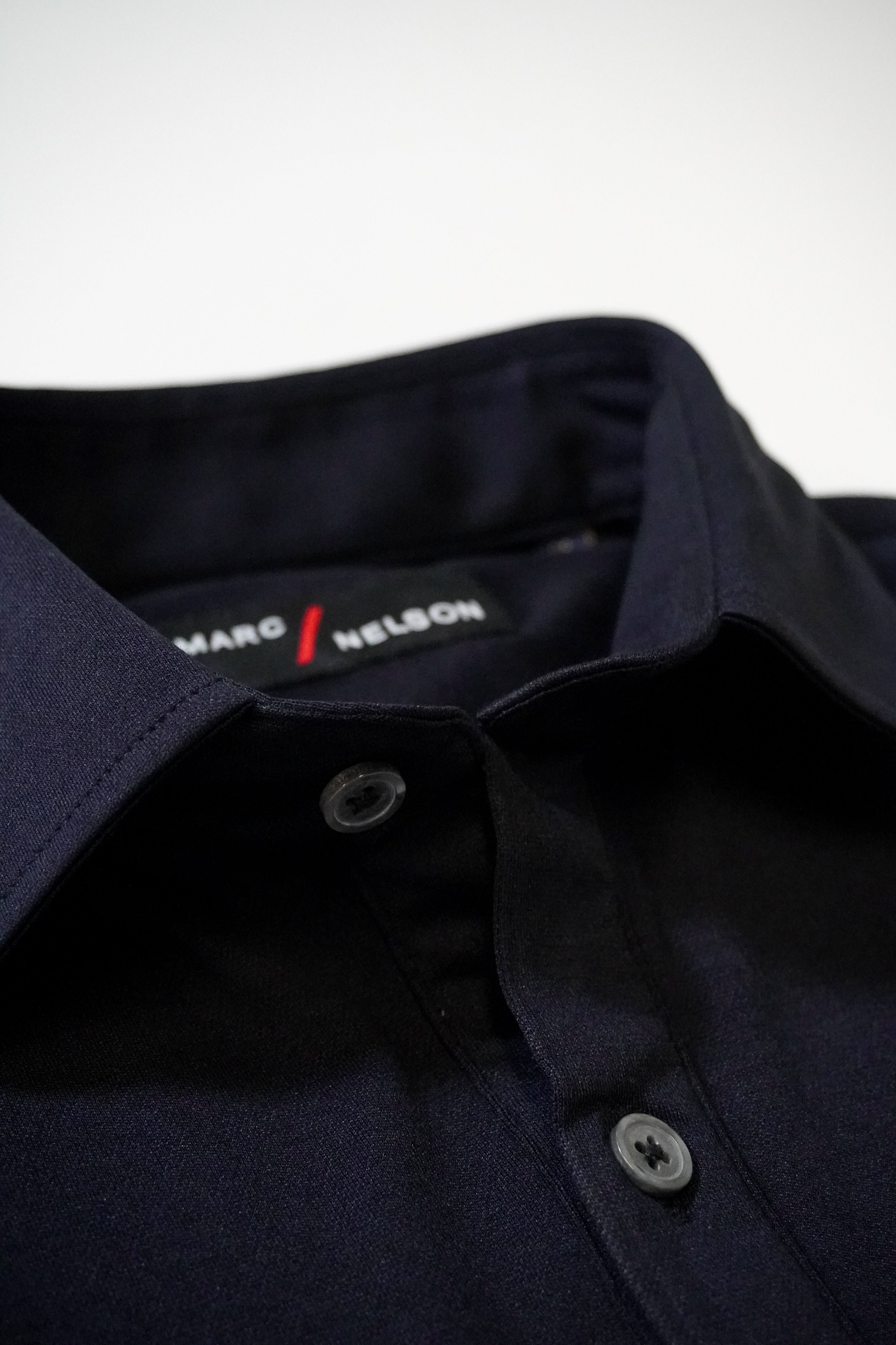 Close-up of a neatly folded dark navy blue collared shirt with gray buttons. The label inside the collar reads “MARC NELSON.” The background is a plain, light surface.