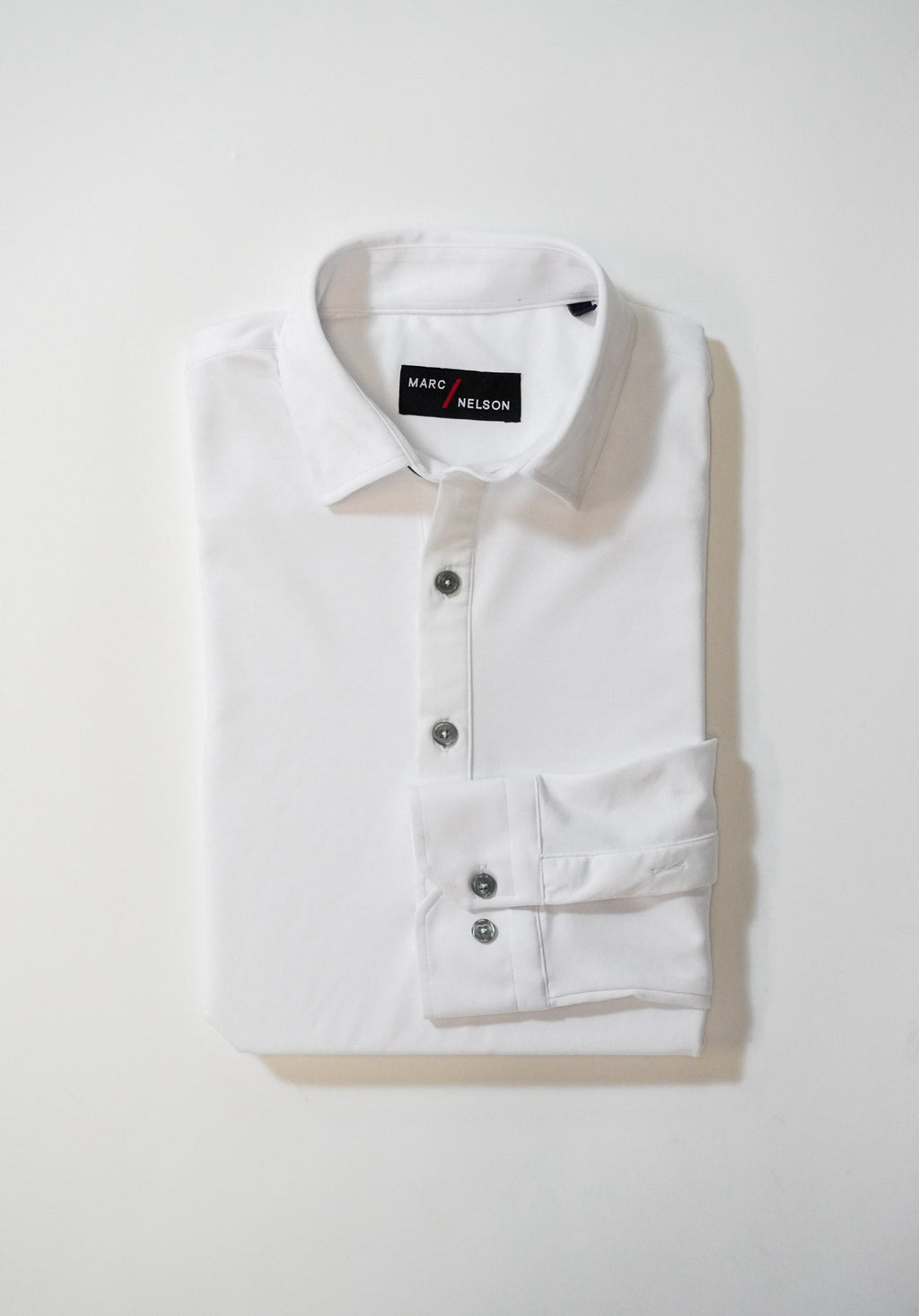 A neatly folded white button-up shirt with a collar and three visible buttons, placed on a white surface. The label inside the collar reads MARC NELSON.