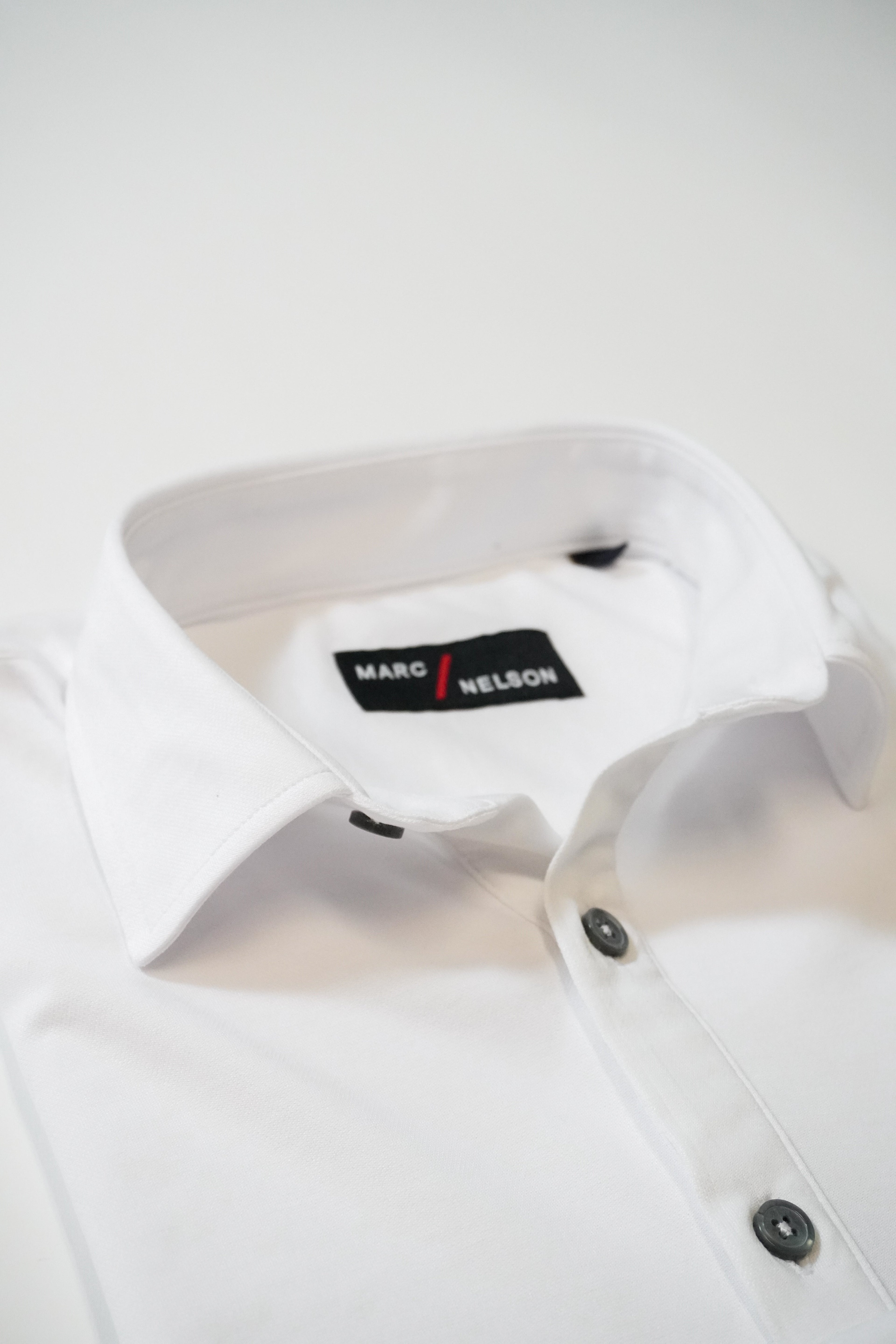 A neatly folded white dress shirt with black buttons, featuring a visible black label on the inside collar that reads MARC / NELSON in white and red text, set against a plain white background.