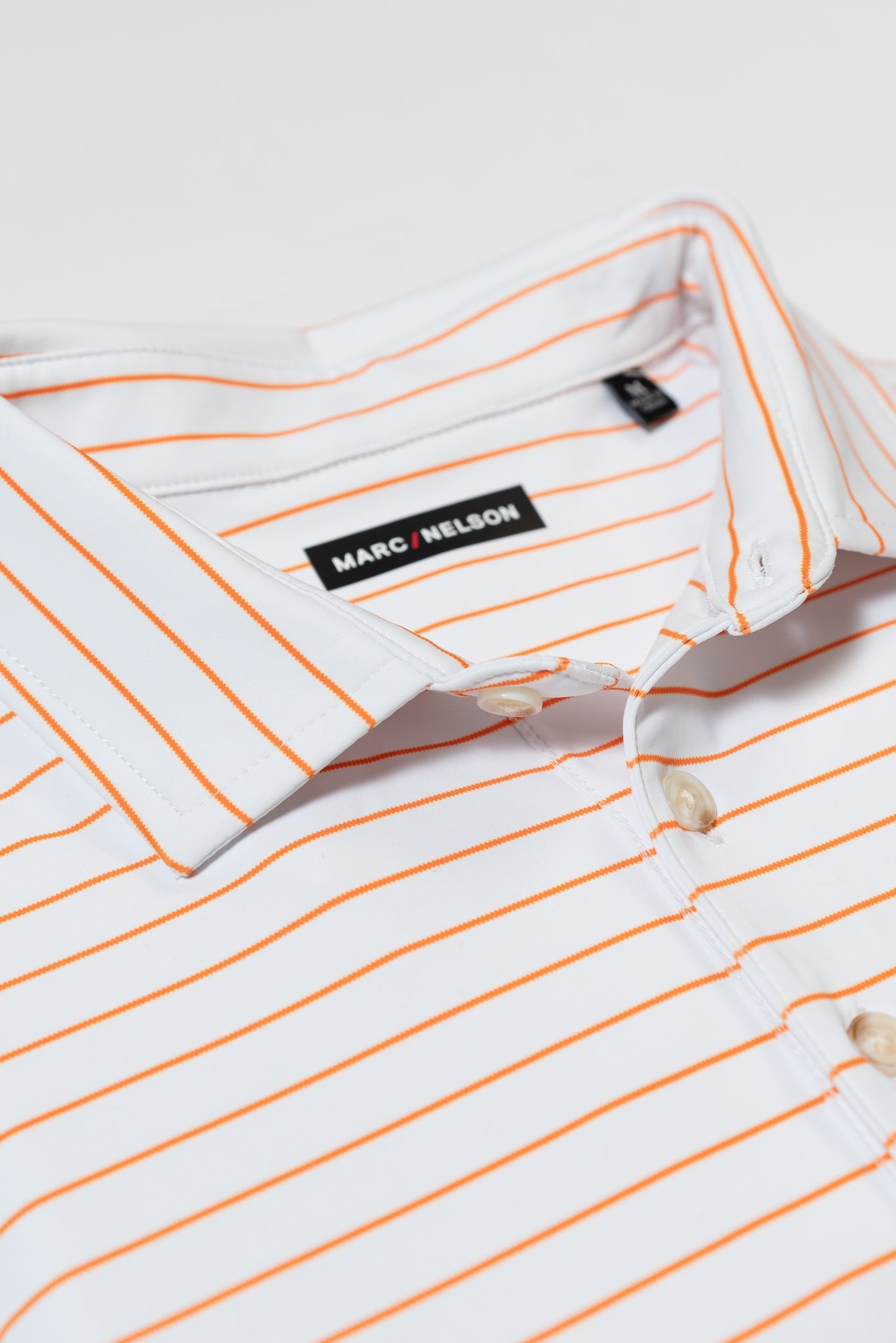 A white collared shirt with thin orange horizontal stripes, featuring a Marc Nelson label on the inside of the collar and white buttons on the front. The shirt is laid flat on a white surface.